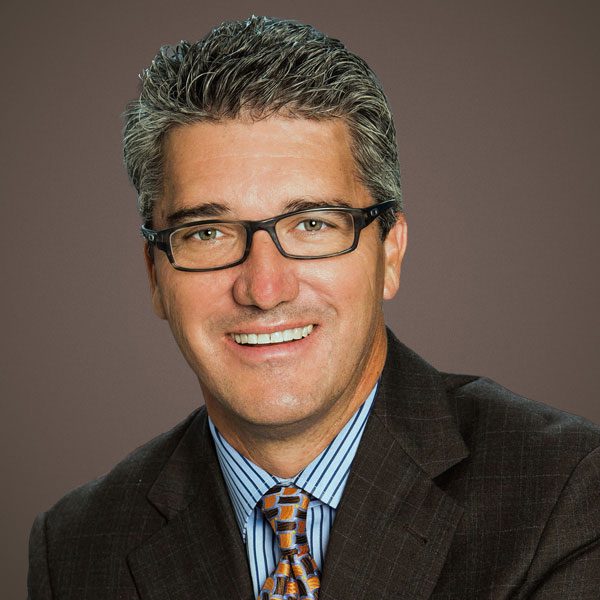 A man wearing glasses, a dark suit, a blue striped shirt, and a patterned tie, smiling in front of a plain brown background.