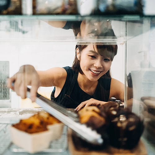 Cheerful Young Asian Woman Working At A Bakery And Serving Fresh Bakery From The Display Cabinet