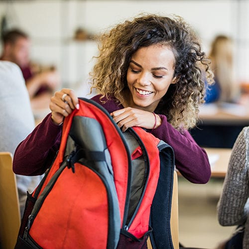 Happy African American Female Student Taking Something From A Backpack In The Classroom.