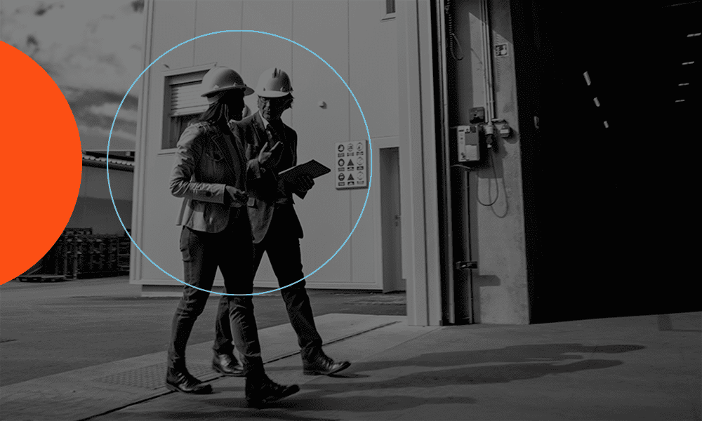 Two people wearing hard hats and business attire walk outside an industrial building, with one holding a tablet. A blue circle and partial orange circle are overlaid on the image.