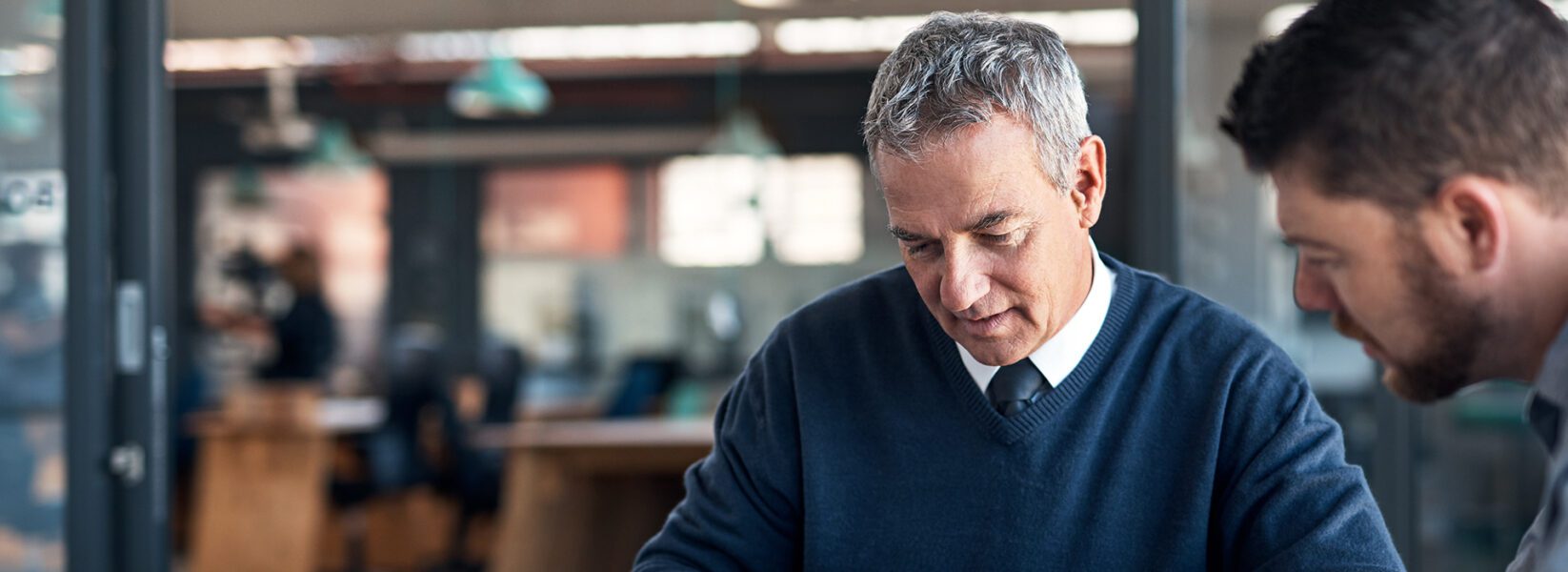 Two men in business attire are sitting at a desk in an office, looking down at documents or paperwork together.