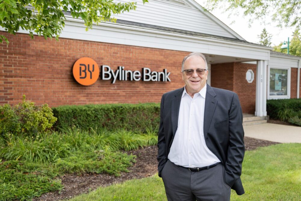A man in a suit stands in front of a brick building with a Byline Bank sign on the wall.