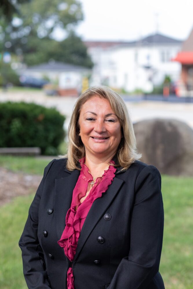 A woman with blonde hair wearing a black blazer and a burgundy blouse stands outdoors, smiling at the camera with a grassy area and buildings in the background.