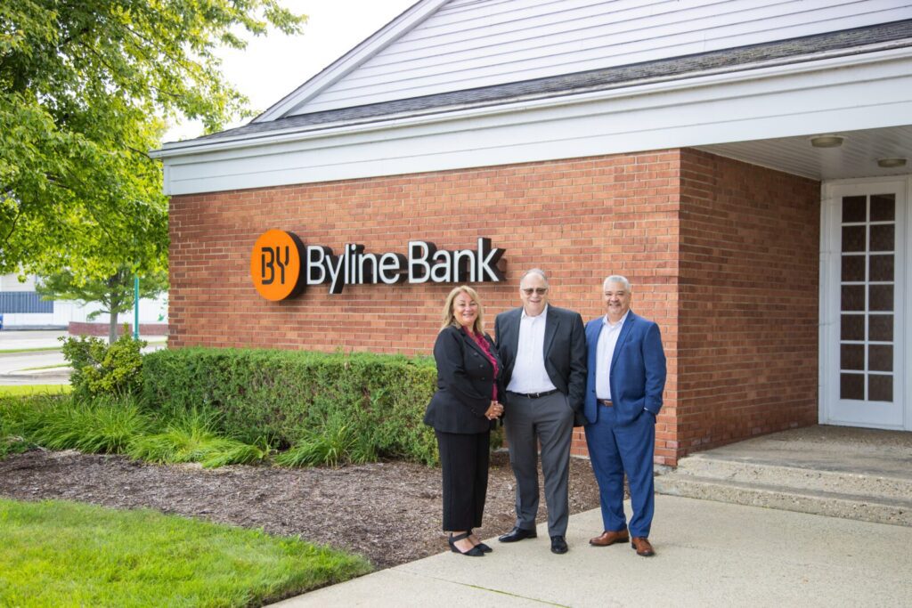 Three people in business attire stand outside a Byline Bank branch building, near a sign with the bank's logo on a brick wall.