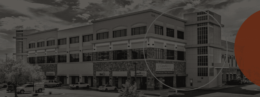 A two-story commercial building with multiple businesses, cars parked in front, and a partly cloudy sky overhead.