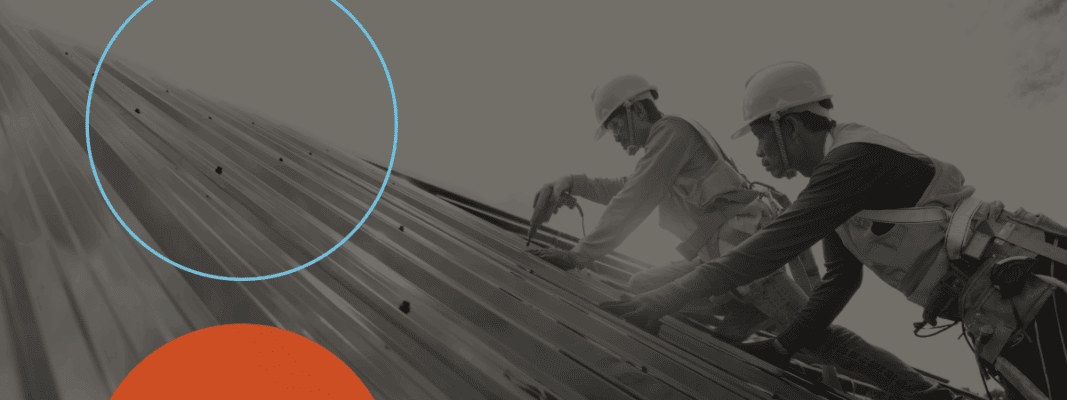 Two construction workers wearing safety gear install metal roofing panels on a sloped roof.