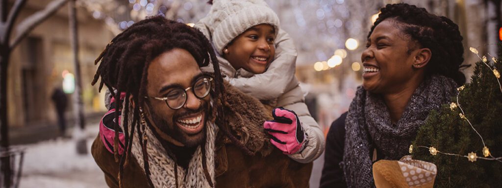 A man gives a young girl a piggyback ride while a woman stands beside them, all smiling outdoors on a winter street decorated with lights.