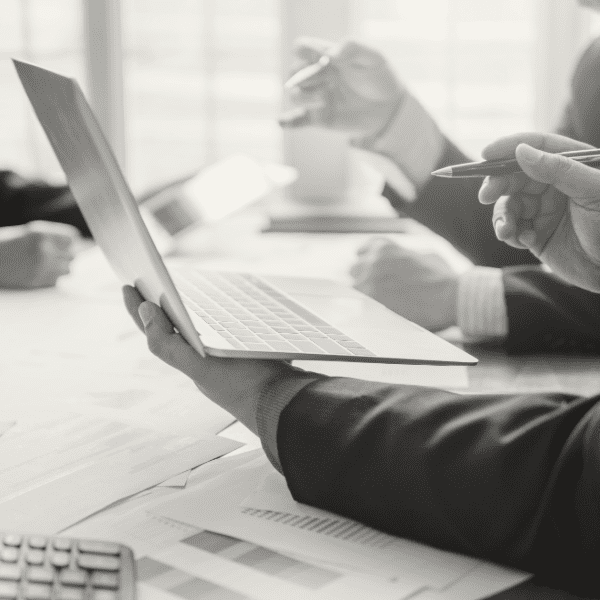 Three people in business attire sit at a table with documents, one holding a laptop and others holding pens, engaged in a meeting discussion.