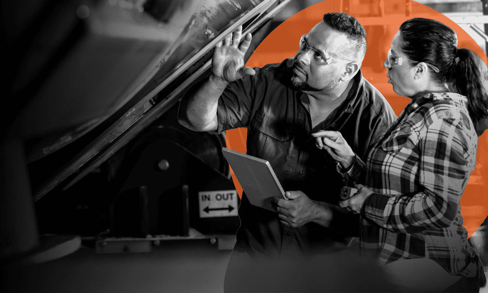 Two people wearing safety glasses examine industrial machinery; one holds a tablet while the other points, with a sign in the background reading "IN OUT.