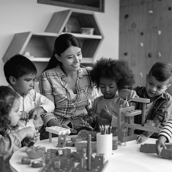 A woman sits at a table with four young children playing with building toys. Shelves and a wall with climbing holds are visible in the background.