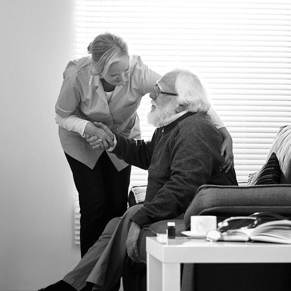 A woman stands beside an elderly man sitting on a couch, shaking his hand and smiling, with a table and books in the foreground.