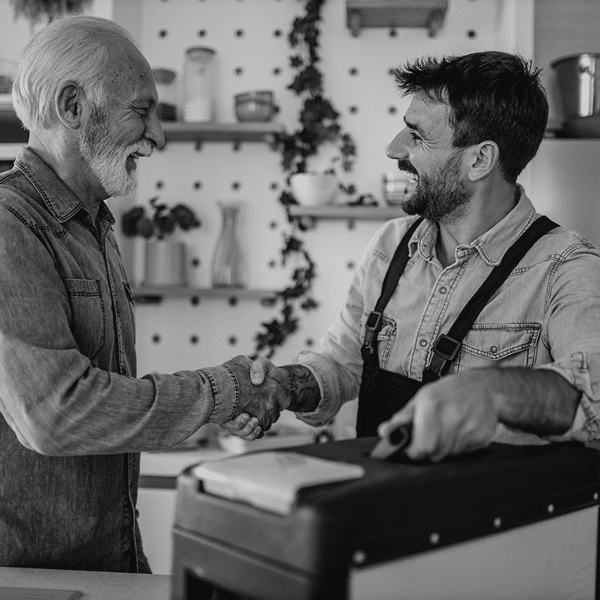 Two men smiling and shaking hands in a kitchen, one wearing an apron and holding a toolbox, suggesting a friendly interaction or agreement.