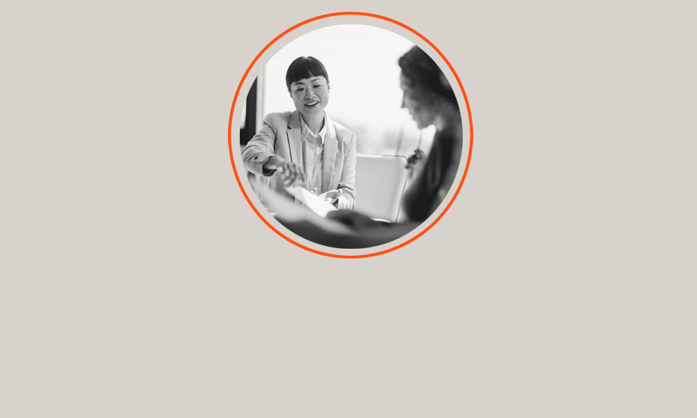 Two people sit at a table, one pointing at a document while the other listens. The photo is black and white and enclosed in a circular frame with an orange border.