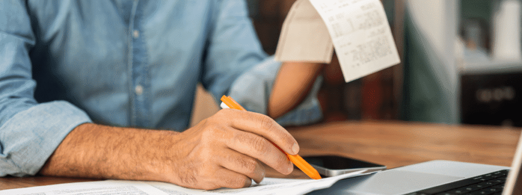 A person reviews receipts and documents while using a laptop, holding a pencil and a phone on the desk.