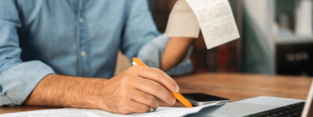 A person reviews receipts and documents while using a laptop, holding a pencil and a phone on the desk.