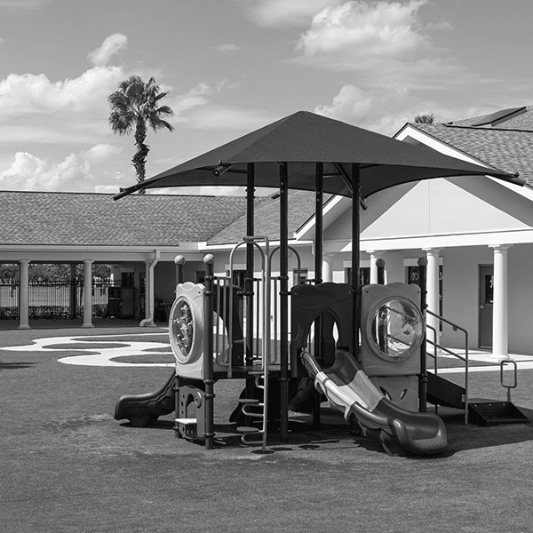 A playground structure with slides and climbing features sits on grass near a building with columns; a palm tree is visible in the background.