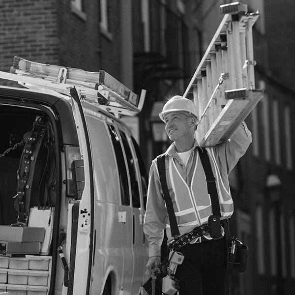 A worker wearing a hard hat and reflective vest carries a ladder on his shoulder while standing next to a work van in an urban area.