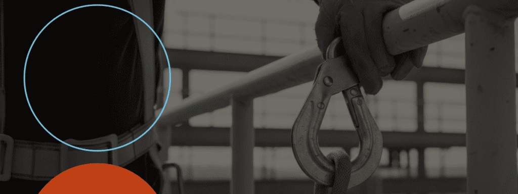 A gloved hand holding a metal safety hook attached to a cable, with building scaffolding visible in the background.