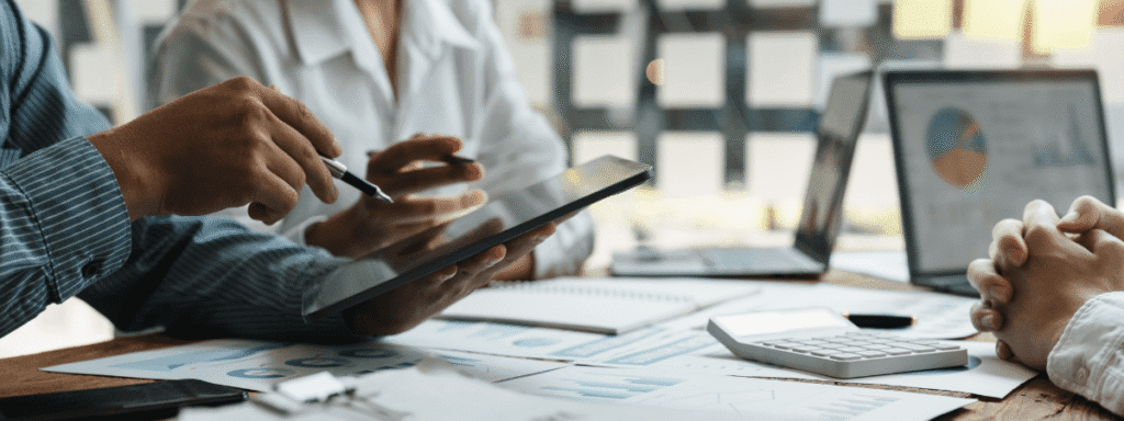 Three people hold a meeting at a table with charts, a calculator, a tablet, and a laptop displaying a pie chart.