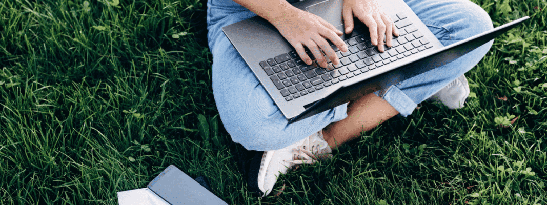 Person sitting on grass, typing on a laptop, with a smartphone and a notebook nearby.