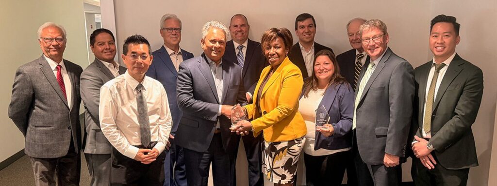 A group of thirteen people pose indoors, with two people in the center shaking hands and holding awards. Most are smiling and dressed in business attire.