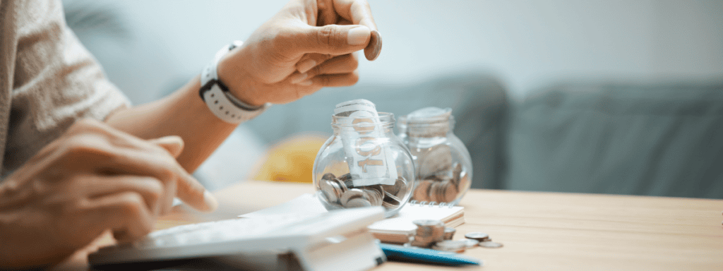 A person counts coins at a table with a calculator, a notebook, and two jars filled with coins, suggesting budgeting or saving money.