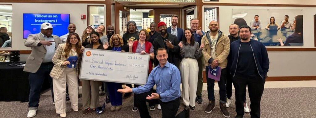 A group of people poses indoors, some smiling and holding a large check labeled “Social Impact Initiative” for $1,000. A man kneels in front, and a digital screen is visible in the background.