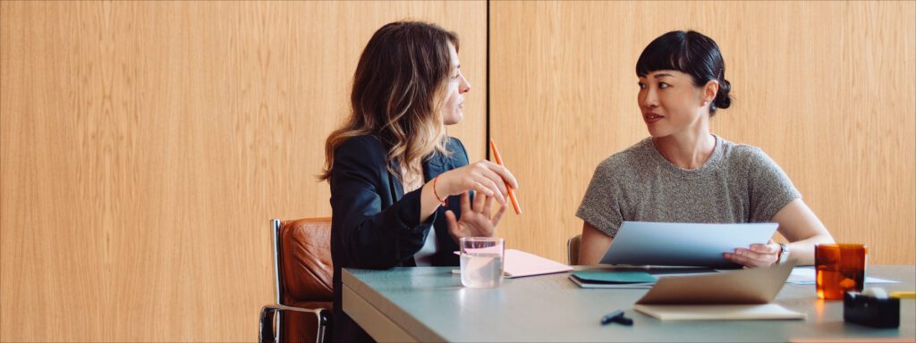 Two women sit at a table having a discussion. One holds a pen and notebook, the other holds papers. Laptops and glasses of water are on the table, with a wooden wall in the background.