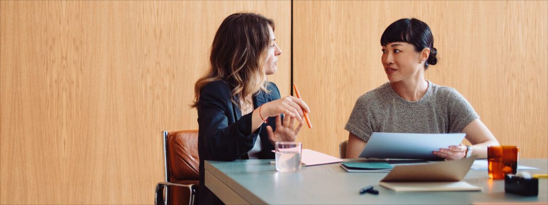 Two women sit at a table having a discussion. One holds a pen and notebook, the other holds papers. Laptops and glasses of water are on the table, with a wooden wall in the background.