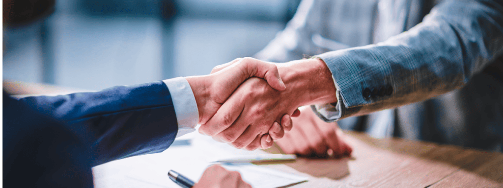 Two people in business attire shaking hands over a desk with documents.