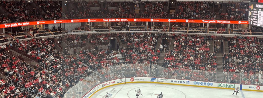 High-angle view of a crowded hockey arena with a digital scoreboard along the mezzanine and a game in progress on the ice.